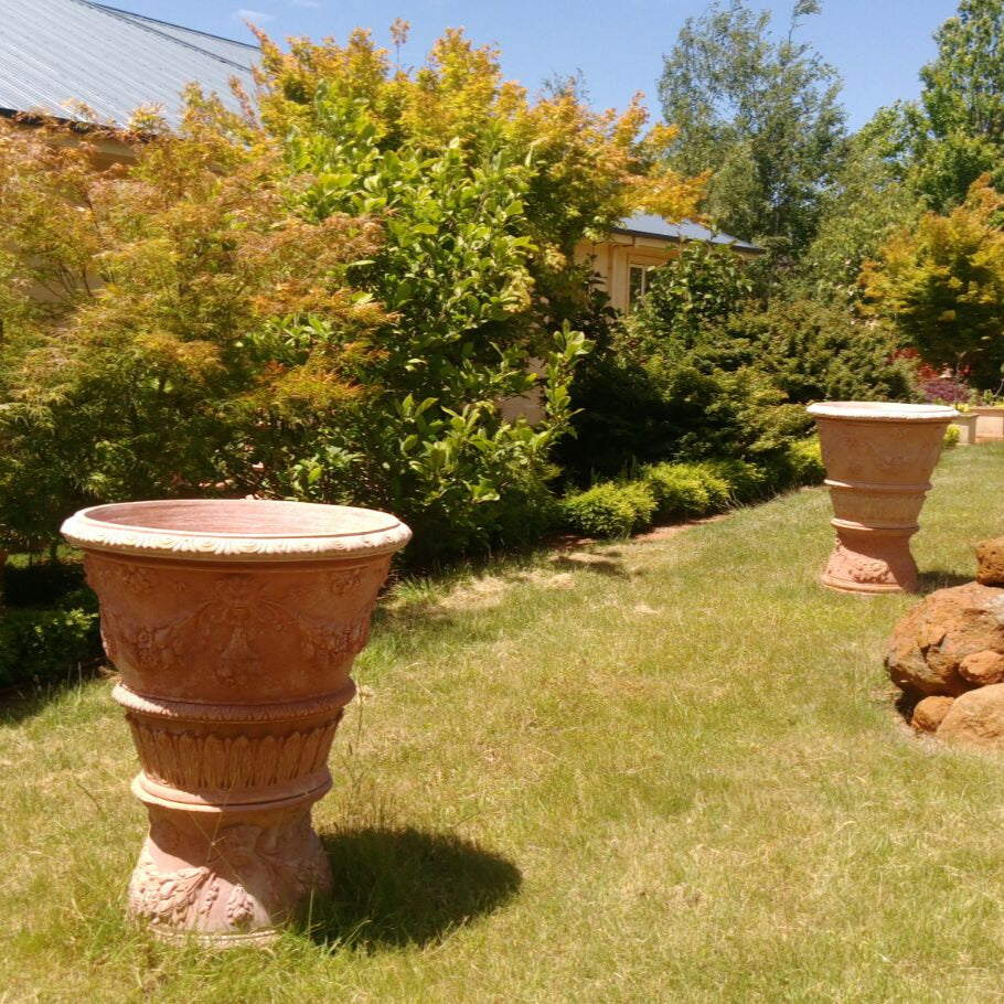 Pair of Vaso Ornato Regale Italian terracotta planters on pedestals placed on lawn, mixed deciduous garden border and outbuilding in background