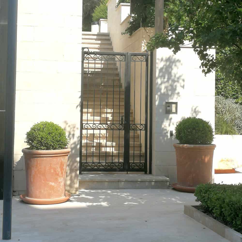 Pair of Vaso Tamburo Liscio Italian terracotta planters with clipped boxwood balls flanking wrought iron garden gate, limestone facade and exterior stair in background